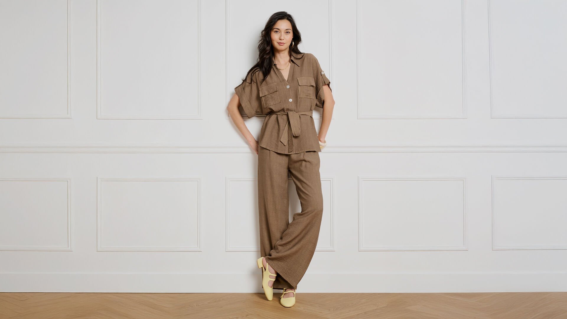 Woman in a brown belted top and wide-leg trousers standing against a white paneled wall.