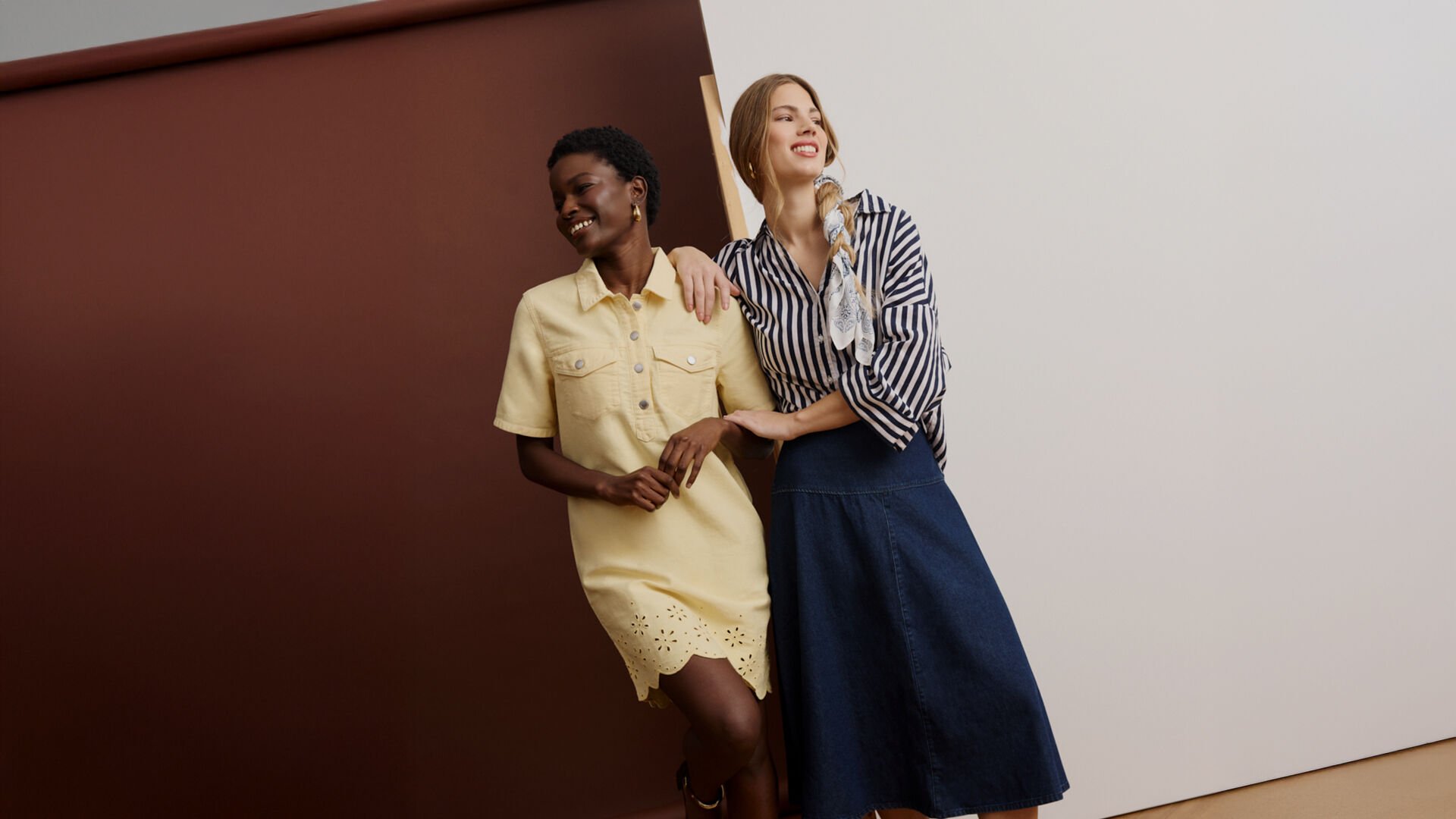 Two women standing together, one in a yellow shirt dress and the other in a striped blouse with a denim skirt.