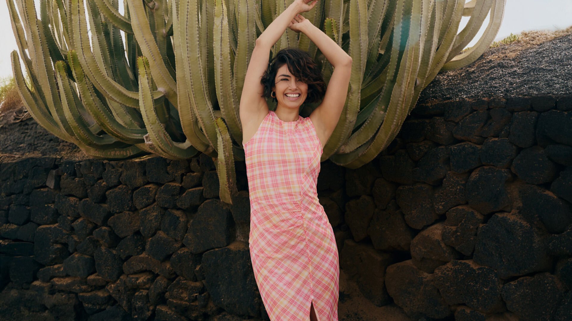 A smiling person with short, dark wavy hair stands with their arms raised in front of a large, multi-armed cactus and a dark stone wall. She are wearing a pink and yellow plaid sleeveless dress.